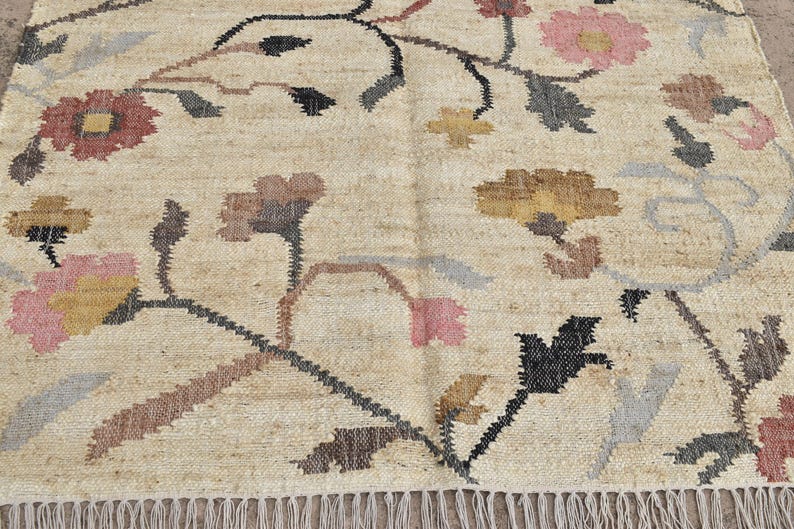 May include: Beige rug with floral design. The rug features a repeating pattern of flowers in shades of pink, brown, yellow, and gray, with black stems and leaves. The rug has a fringed edge.