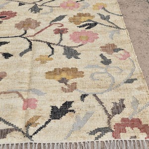 May include: Beige floral rug with a repeating pattern of flowers in pink, brown, and yellow. The rug has a fringe along the bottom edge and is made of a woven material.