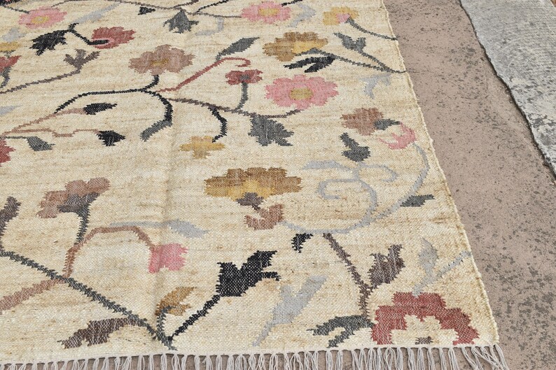 May include: Beige floral rug with a repeating pattern of flowers in pink, brown, and yellow. The rug has a fringe along the bottom edge and is made of a woven material.