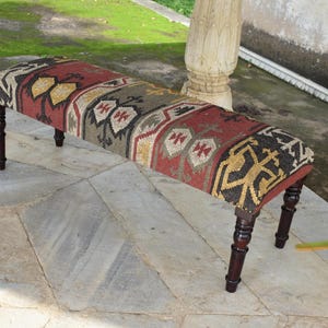 Puede incluir: Banco rectangular con asiento textil estampado en tonos rojos, dorados y negros. El banco tiene cuatro patas de madera torneada de color marrón oscuro. El asiento presenta un diseño geométrico.