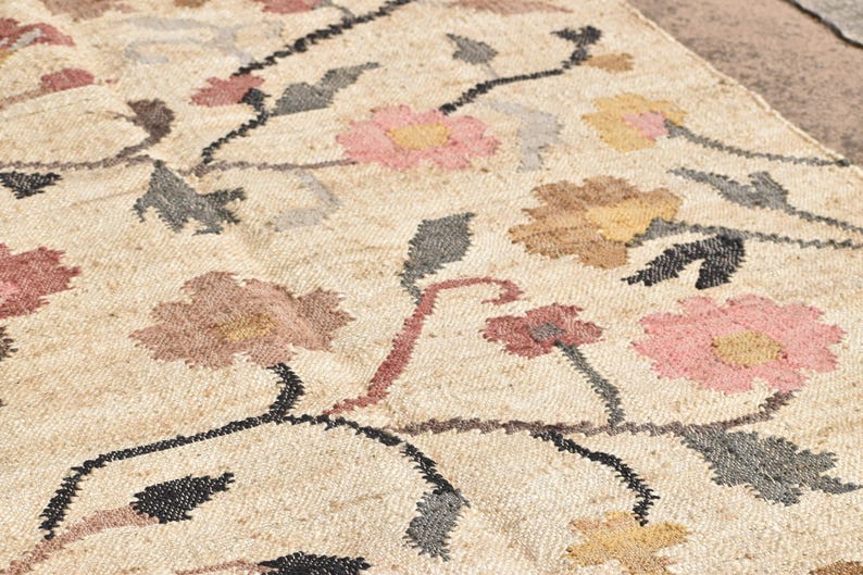 May include: Close-up of a beige rug with a floral pattern. The rug features flowers in shades of pink, brown, and yellow, with black stems and gray leaves. The rug has a textured appearance.