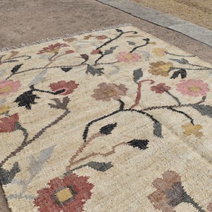 May include: Beige floral rug with a repeating pattern of pink, red, yellow, and brown flowers. The rug has a woven texture and a fringe along one edge. The floral design is set against a neutral background.