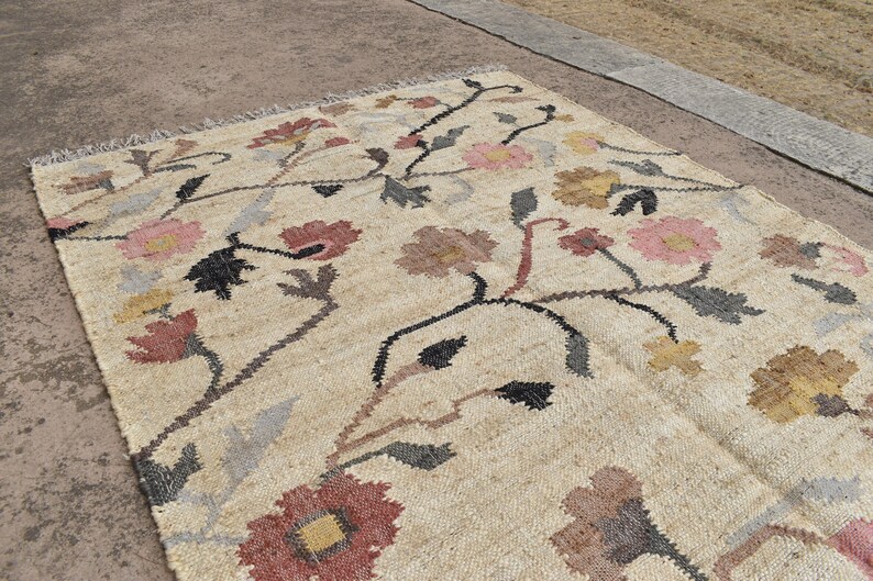 May include: Beige floral rug with a repeating pattern of pink, red, yellow, and brown flowers. The rug has a woven texture and a fringe along one edge. The floral design is set against a neutral background.