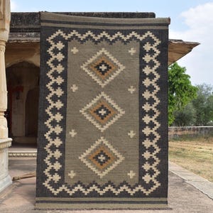 May include: A rectangular rug with an olive green central field, framed by a dark grey border with a zigzag pattern. The rug features three diamond motifs with black, white, and gold accents. The rug is displayed outdoors.