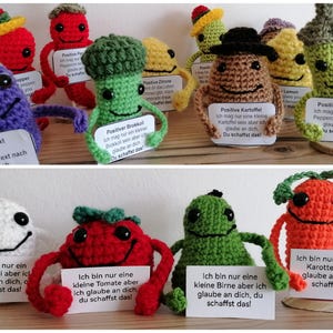 Op de afbeelding: Een verzameling kleurrijke gehaakte figuren, elk representatief voor een ander fruit of groente. Elk figuur houdt een bord vast met een positieve boodschap en de tekst "You schaffst das!" wat in het Nederlands "Je kunt het!" betekent.