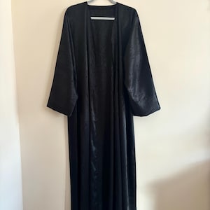 Peut inclure: Longue robe noire à devant ouvert avec un motif subtil texturé, suspendue à un cintre gris contre un mur neutre. La robe a des manches longues et une silhouette fluide, idéale pour superposer.