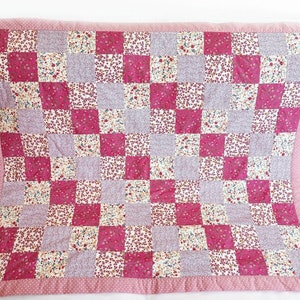 Puede incluir: Una colcha de retazos rosa y blanca con un patrón floral. La colcha está hecha de cuadrados de diferentes telas, y los bordes están recortados con un borde rosa.