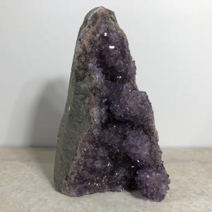 May include: A vertical amethyst geode specimen featuring a cluster of deep purple, crystalline points on one side and a solid, rough, grey-toned rock exterior on the other. The crystalline structure displays varied shades of violet and translucent facets.