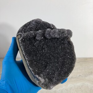 May include: A dark purple amethyst geode specimen featuring a dense cluster of small, sparkling crystals covering the interior surface. The geode has a rounded, irregular shape with a natural grey stone exterior edge visible along the side.