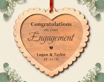 Op de afbeelding: Een houten, hartvormig verlovingsornament met geschulpte randen. Het ornament bevat de tekst "Gefeliciteerd met je verloving", de namen "Logan & Taylor" en de datum 23-11-25. Een rood lint is bevestigd om op te hangen.