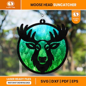 以下が含まれることがあります： 黒いヘラジカの頭のサンキャッチャー。背景は森の風景。ヘラジカの頭はシルエットで、詳細な角があります。背景は青と緑のグラデーションで、森のデザインです。「MOOSE HEAD SUN CATCHER」の文字が上部にあります。