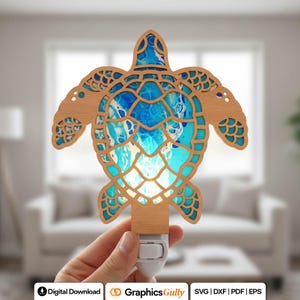 May include: Wooden turtle night light with a blue and white abstract design. The turtle's shell is filled with a vibrant, ocean-inspired pattern. The night light has a plug-in base and is held by a hand. The design is laser cut.
