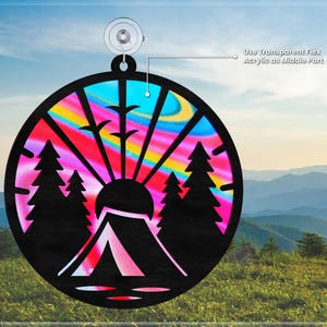 May include: A round, colorful, wooden ornament with a silhouette of a tent, trees, and a sun. The ornament has a rainbow swirl pattern and is designed to be hung.