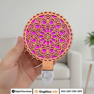 Puede incluir: Una luz de noche decorativa con un diseño de mandala de madera cortada con láser y un fondo rosa vibrante. El intrincado patrón está enmarcado en un marco redondo y festoneado. La luz de noche tiene una base blanca y un enchufe de madera.