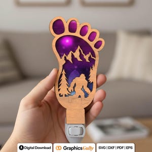 May include: A wooden night light shaped like a foot, featuring a Bigfoot silhouette in a mountain and forest scene. The interior glows with purple and blue light. The night light has a standard plug.