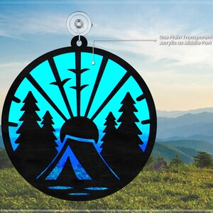 May include: A round, blue and black ornament with a silhouette of a tent, trees, and a sun rising over a mountain. The ornament is designed to be layered with a clear acrylic piece in the center.