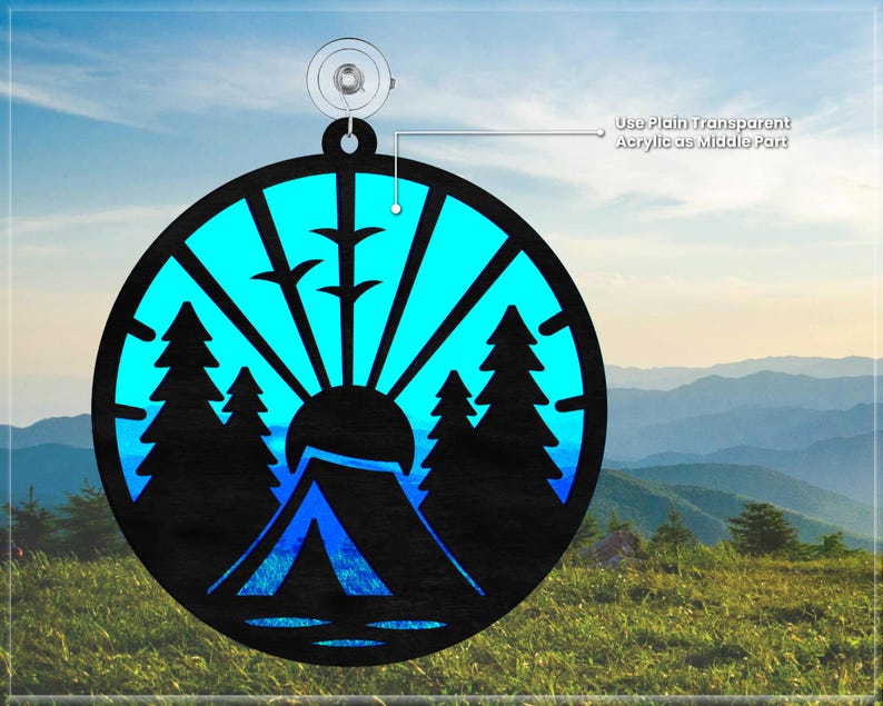 May include: A round, blue and black ornament with a silhouette of a tent, trees, and a sun rising over a mountain. The ornament is designed to be layered with a clear acrylic piece in the center.