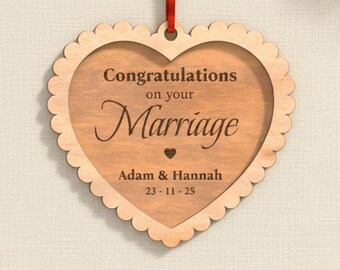Op de afbeelding: Houten hartvormige huwelijksdecoratie met geschulpte randen. De tekst "Congratulations on your Marriage" is in het midden gegraveerd, samen met de namen "Adam & Hannah" en de datum "23-11-25". Een rood lint is bevestigd om op te hangen.