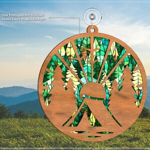 May include: A wooden ornament shaped like a circle with a stained glass design of a forest scene. The ornament features a tent, trees, and a sun. The ornament is hanging from a suction cup.