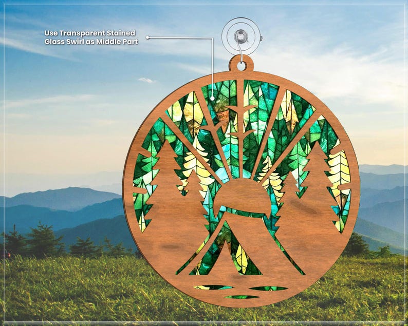 May include: A wooden ornament shaped like a circle with a stained glass design of a forest scene. The ornament features a tent, trees, and a sun. The ornament is hanging from a suction cup.