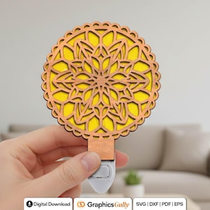 May include: A decorative night light with a laser-cut wooden design. The night light features a circular, floral pattern with a yellow background. The wooden frame is a warm brown color. The night light is plugged into a white base.