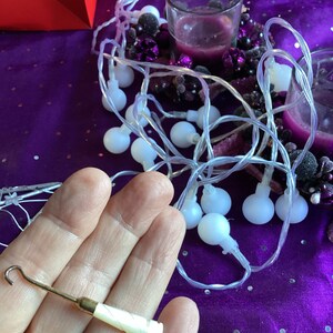 Peut inclure: Un petit crochet ancien avec un manche blanc et une tige en métal doré. Le crochet est présenté sur un fond violet avec des guirlandes lumineuses et un bougeoir. C'est un outil de couture vintage.