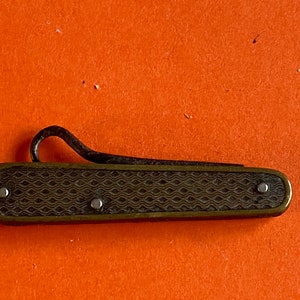 May include: A small, antique pocket knife with a decorative, wave-patterned handle. The handle is a dark brass color, and the blade is folded into the handle. The knife has a curved, hook-shaped element at the top.