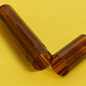 Op de afbeelding: Een cilindrische houten container met een deksel, met een rijk, gestreept patroon van bruine en bruingeel tinten. Het deksel is losgemaakt en onthult de holle binnenkant. De container staat tegen een gele achtergrond.