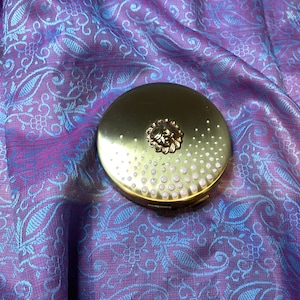 Op de afbeelding: Een goudkleurige compactpoederdoos met een bloemmotief op het deksel en een stippenpatroon langs de rand. De ronde poederdoos ligt op een paars en blauw patroonweefsel.