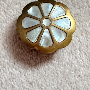 May include: A brass-toned, flower-shaped button with a mother-of-pearl inlay. The button has a scalloped edge and a central circular design. The mother-of-pearl segments are iridescent, reflecting light. The button is likely for clothing or decorative purposes.