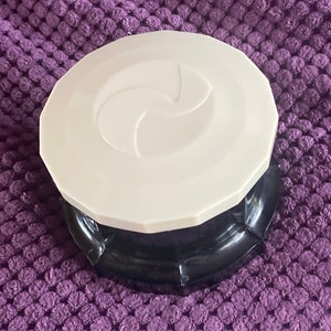 Può includere: Un contenitore rotondo bicolore con un coperchio bianco e una base nera. Il coperchio bianco presenta un disegno a spirale al centro. Il contenitore è su una superficie viola strutturata.