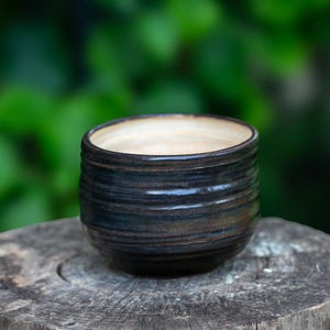 May include: A small, handmade ceramic bowl with a dark blue and brown exterior and a light beige interior. The bowl has horizontal ridges and sits on a weathered wooden surface. The background is a blurred green.