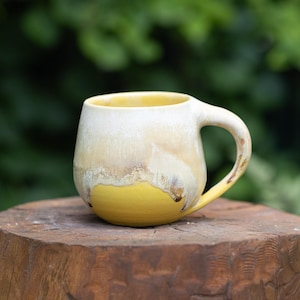 Peut inclure: Une tasse en céramique arrondie présentant un design à deux tons. La partie supérieure est d'un blanc cassé crémeux qui se transforme en une base jaune vif. La tasse possède une anse incurvée et une finition lisse et brillante.