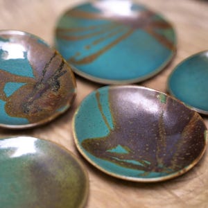 Può includere: Cinque piccole ciotole in ceramica fatte a mano con smalto turchese e marrone. Le ciotole hanno forme organiche e motivi diversi, con finitura lucida. Le ciotole sono disposte su una superficie di legno, mostrando le loro trame e colori unici.