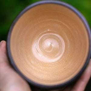 May include: A handmade ceramic bowl with a light brown interior and a dark brown rim. The bowl is held in a person's hand, with a blurred green background. The bowl's interior has a spiral pattern.