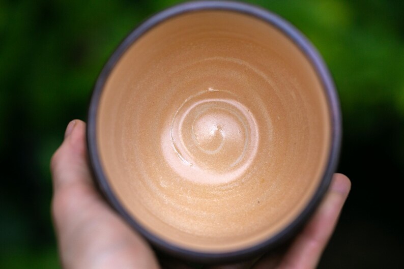 May include: A handmade ceramic bowl with a light brown interior and a dark brown rim. The bowl is held in a person's hand, with a blurred green background. The bowl's interior has a spiral pattern.
