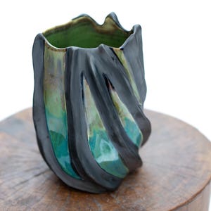 May include: A handcrafted ceramic vase with a unique, textured design. The vase features a dark grey exterior with vertical, wave-like patterns that reveal shades of teal and green. The interior is a vibrant green. The vase sits on a wooden surface.