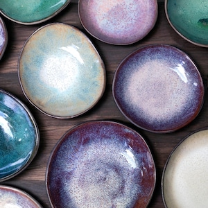 Könnte beinhalten: Draufsicht auf eine Sammlung von Keramikplatten in verschiedenen Farben, darunter Türkis, Lila und Creme. Die Teller haben unregelmäßige Formen und glänzende Oberflächen, mit einem dunkelbraunen Holzuntergrund.