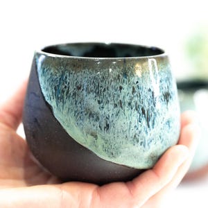 Pu&ograve; includere: Una tazza in ceramica fatta a mano di forma arrotondata. La tazza ha una base marrone scuro che sfuma in una glassa turchese e blu nella met&agrave; superiore. La tazza &egrave; tenuta in una mano.