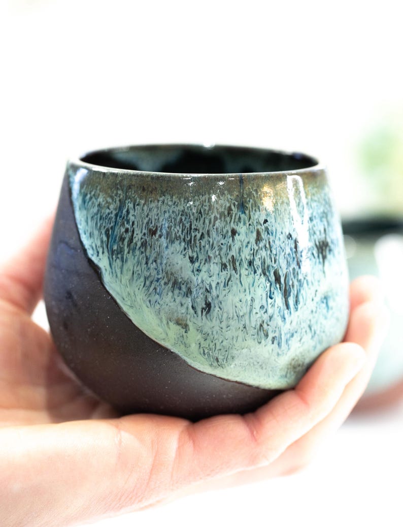 Pu&ograve; includere: Una tazza in ceramica fatta a mano di forma arrotondata. La tazza ha una base marrone scuro che sfuma in una glassa turchese e blu nella met&agrave; superiore. La tazza &egrave; tenuta in una mano.