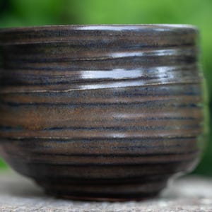 May include: A handcrafted ceramic bowl with a dark brown and blue glaze. The bowl has a rounded shape with horizontal textured lines. The exterior has a glossy finish. The bowl is likely used for serving food or as a decorative item.
