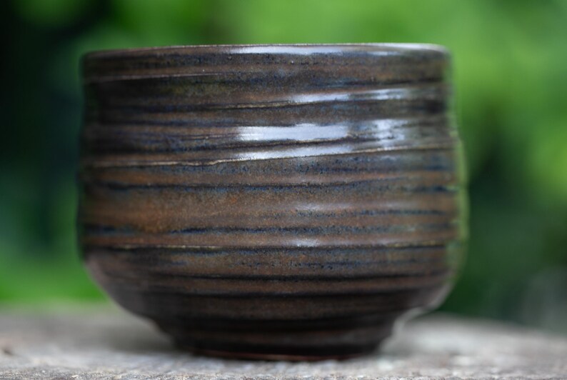 May include: A handcrafted ceramic bowl with a dark brown and blue glaze. The bowl has a rounded shape with horizontal textured lines. The exterior has a glossy finish. The bowl is likely used for serving food or as a decorative item.