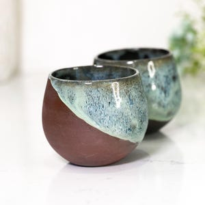 Pu&ograve; includere: Due bicchieri da vino senza stelo in ceramica fatti a mano. La met&agrave; inferiore di ogni bicchiere &egrave; di un marrone rossiccio intenso, mentre la met&agrave; superiore passa a una glassa verde acqua e blu. I bicchieri hanno una forma arrotondata.