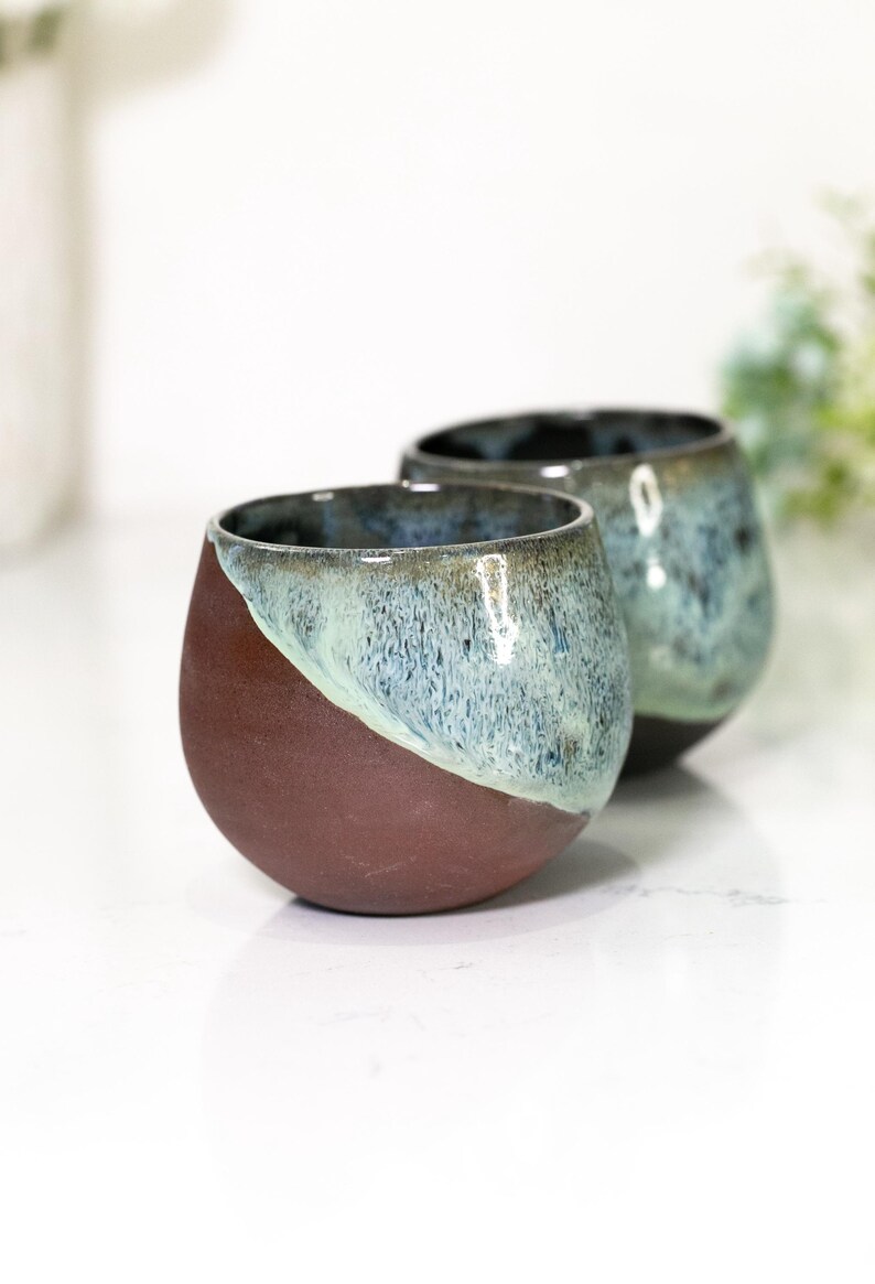 Pu&ograve; includere: Due bicchieri da vino senza stelo in ceramica fatti a mano. La met&agrave; inferiore di ogni bicchiere &egrave; di un marrone rossiccio intenso, mentre la met&agrave; superiore passa a una glassa verde acqua e blu. I bicchieri hanno una forma arrotondata.