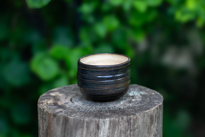 May include: A small, handmade ceramic bowl with a dark blue and brown exterior and a light beige interior. The bowl has horizontal ridges and sits on a weathered wooden surface. The background is a blurred green.