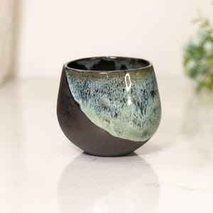Pu&ograve; includere: Una tazza in ceramica fatta a mano con un design unico. La met&agrave; inferiore &egrave; nera opaca, mentre la met&agrave; superiore presenta una miscela di smalto blu e verde. La tazza ha una forma arrotondata e organica, perfetta per caff&egrave; o t&egrave;.