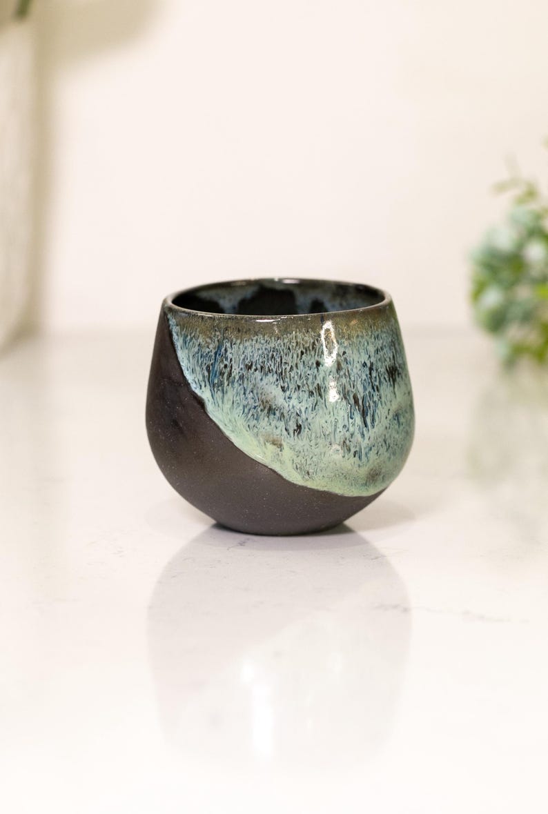 Pu&ograve; includere: Una tazza in ceramica fatta a mano con un design unico. La met&agrave; inferiore &egrave; nera opaca, mentre la met&agrave; superiore presenta una miscela di smalto blu e verde. La tazza ha una forma arrotondata e organica, perfetta per caff&egrave; o t&egrave;.