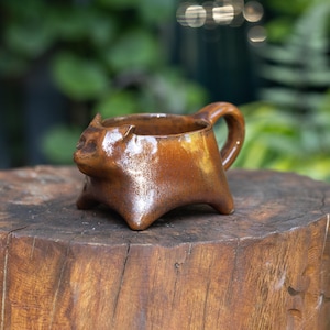 May include: A small, glossy, brown ceramic mug shaped like a cat with pointed ears and a rounded handle. The mug features a smooth, reflective glaze with subtle variations in color, standing on four short, sturdy legs that form the base.