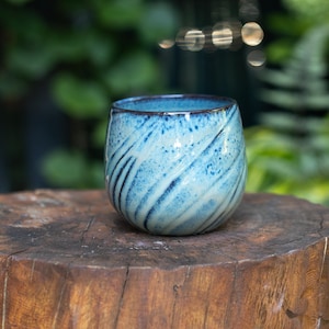 May include: A rounded ceramic cup featuring a speckled light blue and dark blue glaze with a swirling pattern. The rim is dark and defined, contrasting with the lighter body. The cup has a smooth, curved shape without a handle, showcasing a reactive glaze finish that blends various shades of blue across its surface.