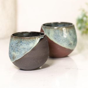 Pu&ograve; includere: Due bicchieri da vino senza stelo in ceramica fatti a mano. La met&agrave; inferiore di ogni bicchiere &egrave; marrone scuro, mentre la met&agrave; superiore &egrave; azzurra e verde. I bicchieri sono su una superficie bianca.